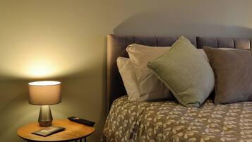 A neatly made bed with patterned bedding, a bedside lamp, a remote control, and a small table with a book.