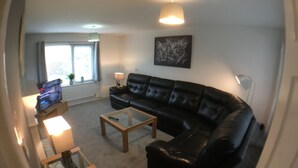 A living room with a black leather sofa, a glass coffee table, a television on a stand, and a window with curtains. - Phoenix House (Corby)