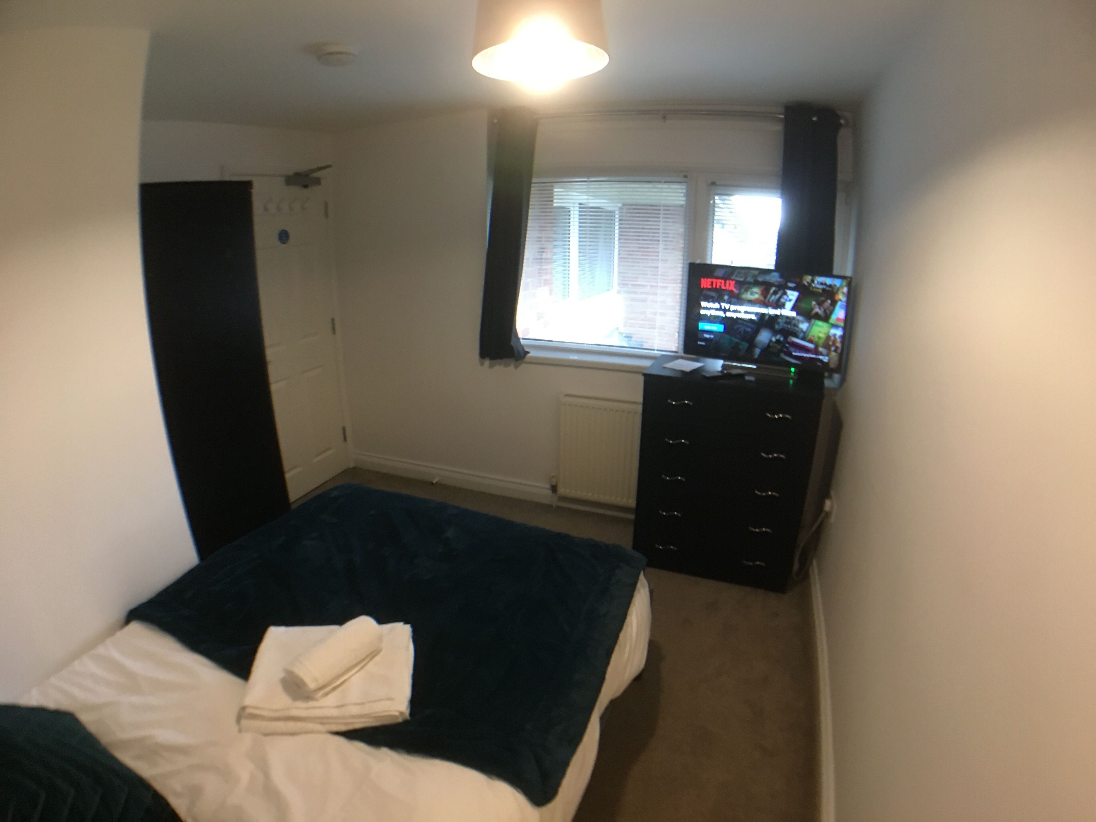 A bedroom with a bed, a dresser, a window with blinds, and a TV displaying Netflix.