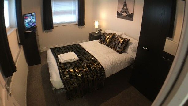 A hotel room with a bed, a nightstand, a lamp, a painting of the Eiffel Tower, a television, and a door. - Phoenix House (Corby)