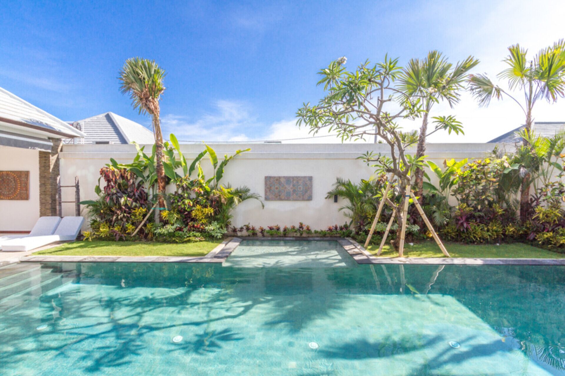 A swimming pool surrounded by lush tropical plants and a white building with a tiled roof.