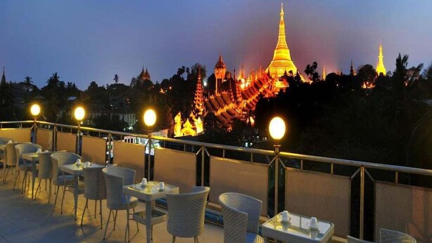 A rooftop terrace with tables and chairs, overlooking a cityscape with illuminated buildings and a prominent golden pagoda. - Hotel Lavender (Yangon)
