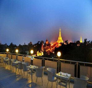 A rooftop terrace with tables and chairs, overlooking a cityscape with illuminated buildings and a prominent golden pagoda. - Hotel Lavender (Yangon)