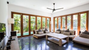 A spacious living room with a large window, a ceiling fan, and a flat-screen TV. - Luxury 5BR Family Villa Near Seminyak, Bali Villa 1167 (Seminyak)