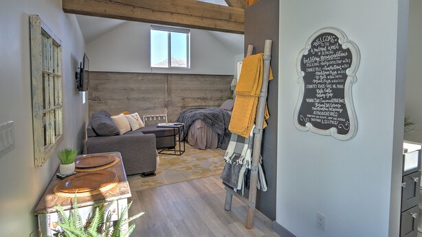 A cozy loft-style room with a loft bed, a wooden ceiling, a chalkboard wall with a welcome message, a small table with plates, and a window providing natural light. - A Night with the Alpacas & Alpaca Experience! (Silt)