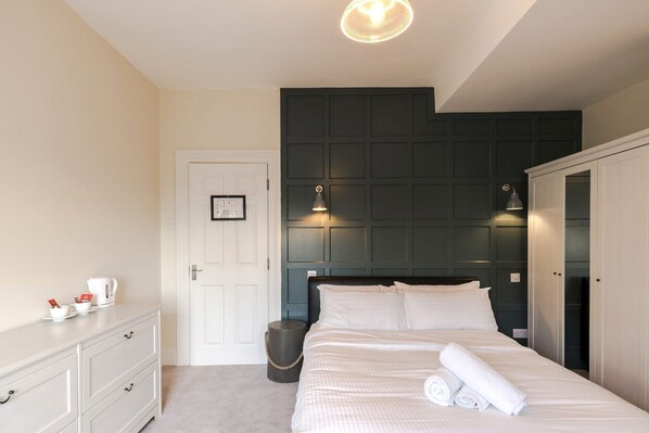 Une chambre à coucher avec un lit, des lampes de chevet, une commode blanche, une porte ornée d’une carte encadrée et un lambris foncé.