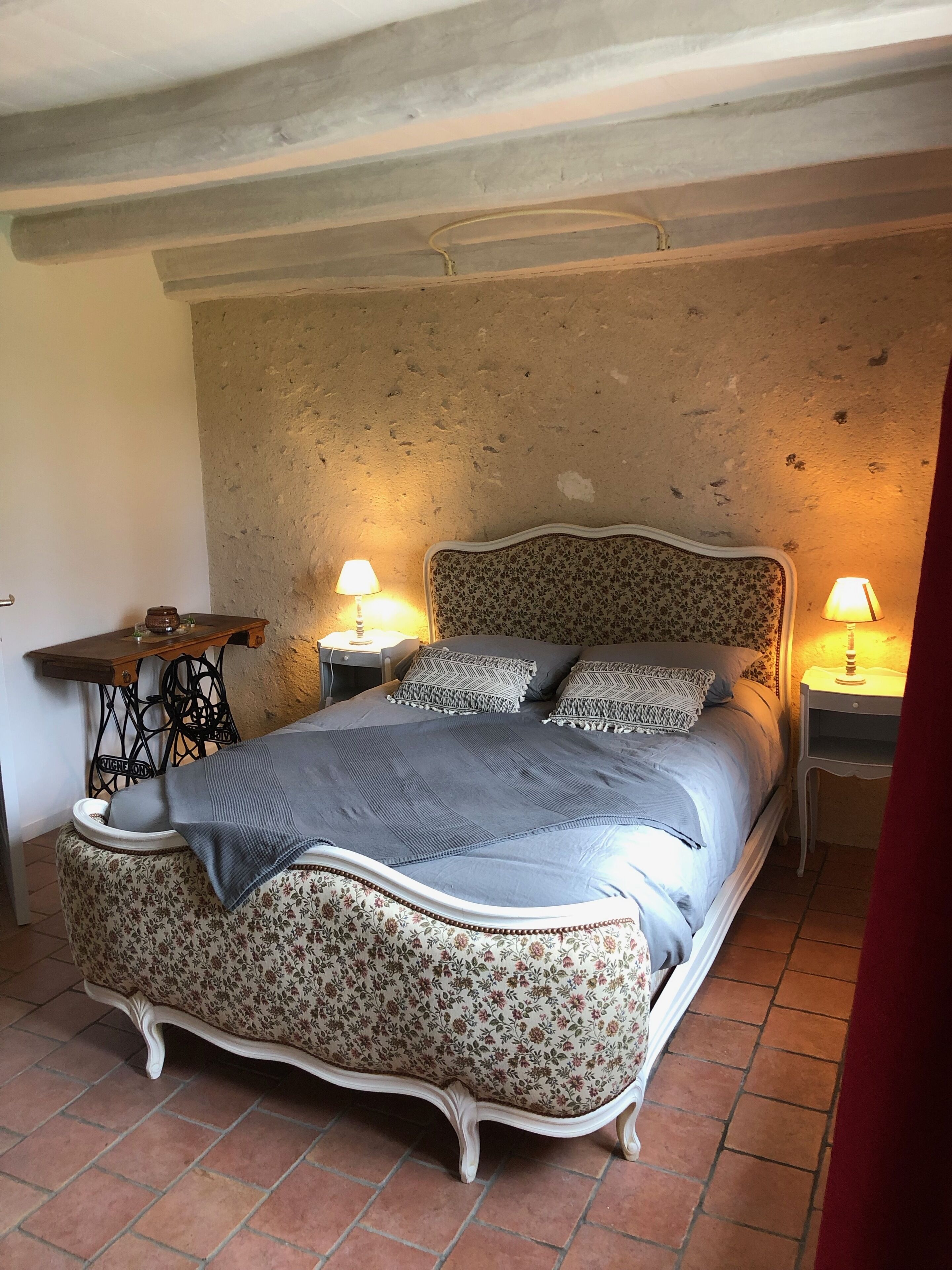Un lit doté d’une tête de lit et d’un pied de lit ornés de motifs floraux, flanqué de deux tables de chevet avec des lampes. La pièce présente un plafond rustique avec des poutres apparentes et un sol carrelé.