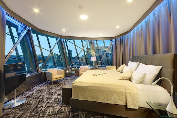 A modern hotel room with a large bed, a sofa, and a chair. The room has large windows offering a view of a bridge and cityscape. - Dancing House Hotel (Prague)