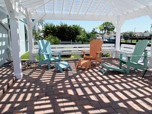 Un patio couvert, agrémenté de chaises Adirondack et d’une table, entouré d’une clôture blanche et de verdure.