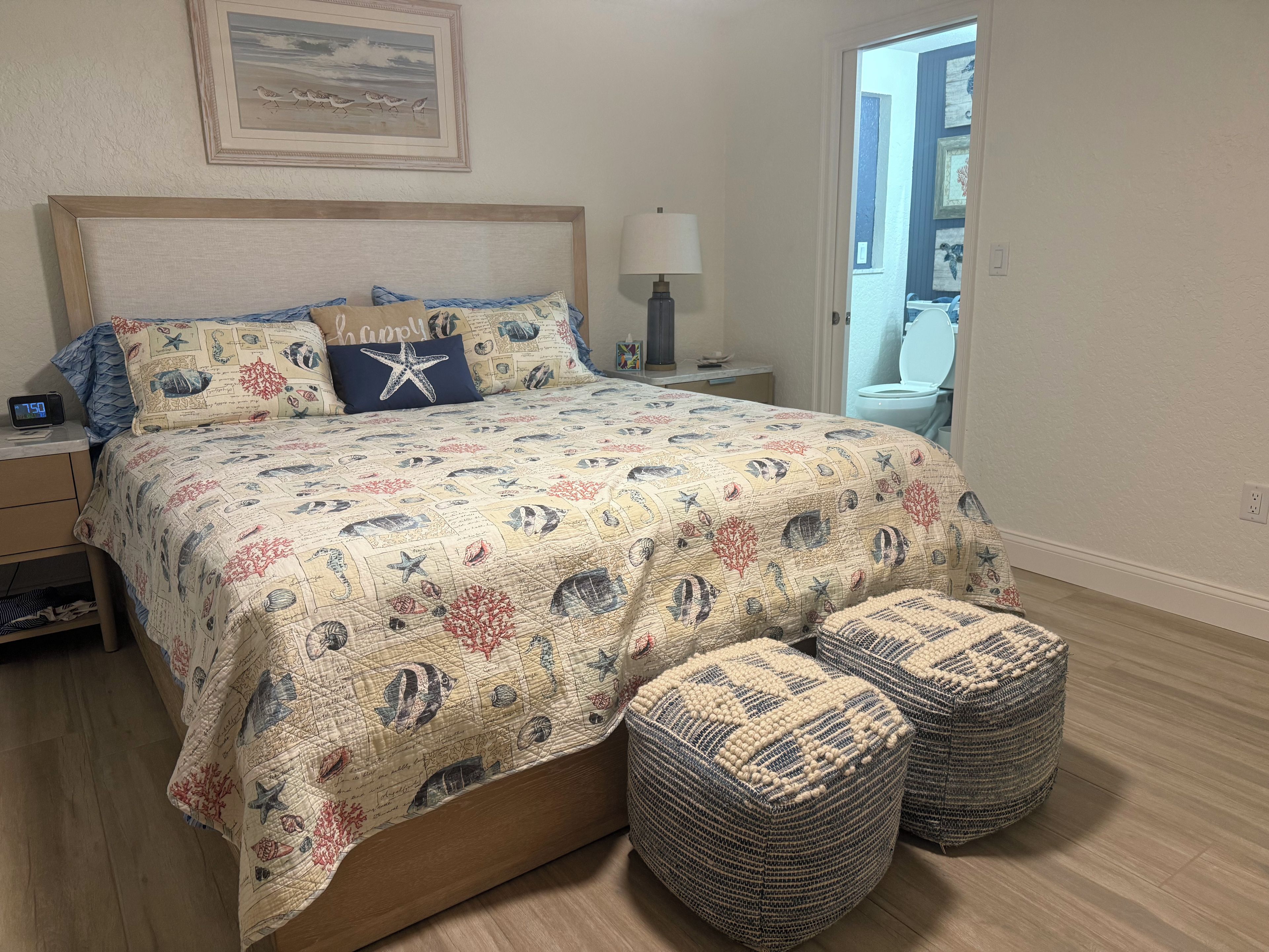 A bedroom with a bed, nightstand, lamp, and two ottomans.