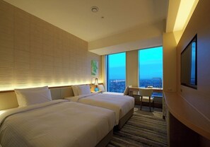 A hotel room with two beds, a desk, a chair, a TV, and a large window with a city view.