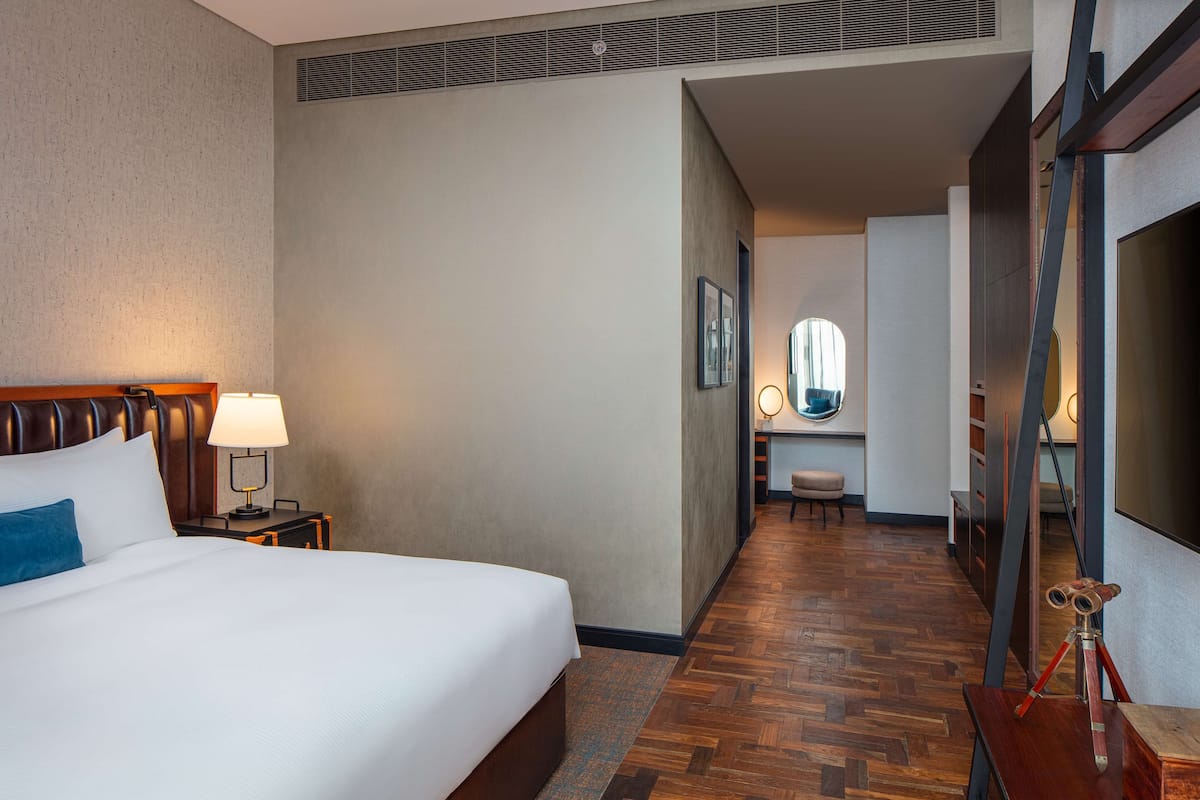 A hotel room with a large bed, a nightstand with a lamp, a wooden floor, a mirror, and a desk with a chair.