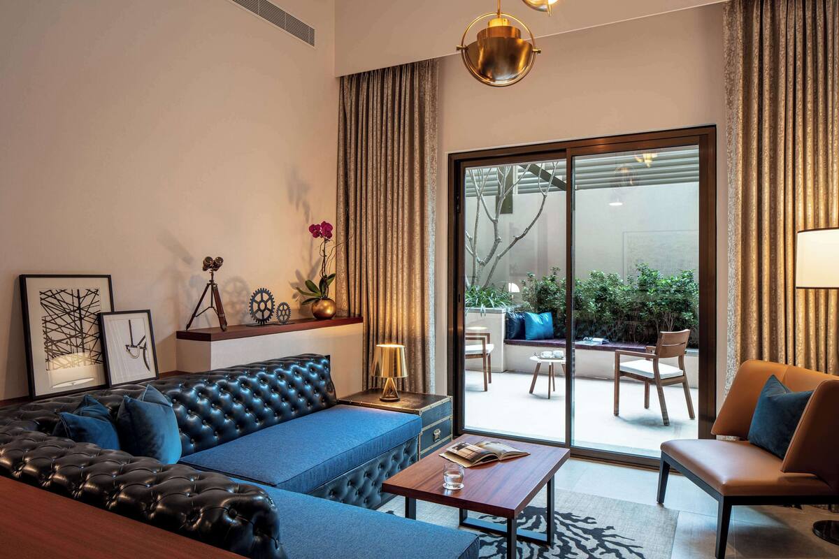 A modern living room with a blue sofa, a leather armchair, a wooden coffee table, and a large sliding glass door leading to an outdoor seating area.