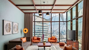 A modern living room with a sofa, two armchairs, a coffee table, and a television. Large windows offer a view of a cityscape.