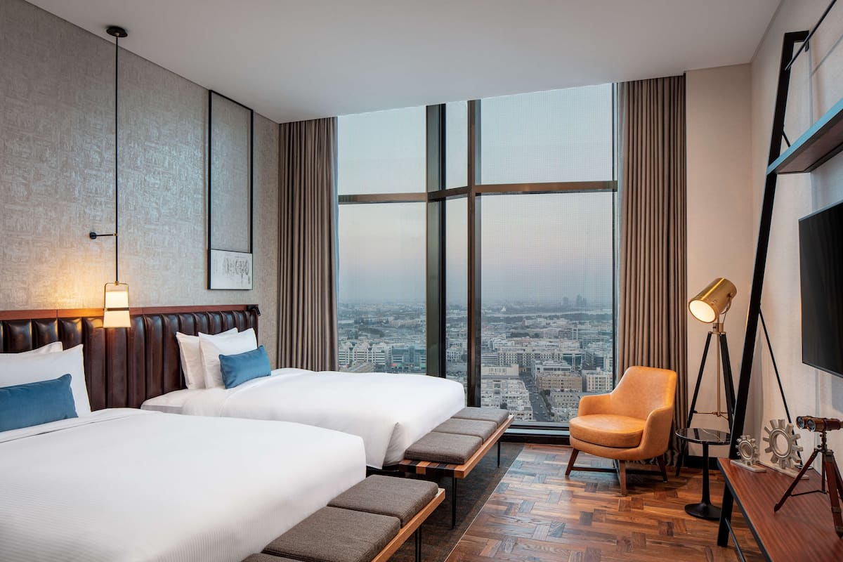 A hotel room with two beds, a view of the city, a large window, a desk with a lamp, and a chair.