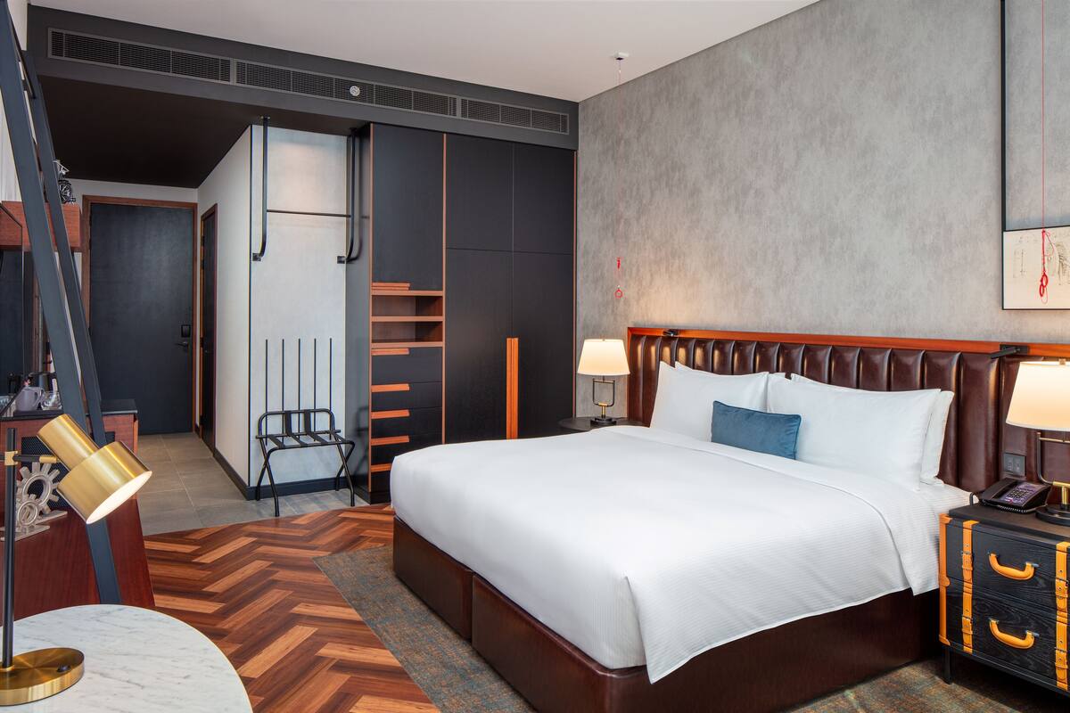 A hotel room with a large bed, bedside lamps, a desk with a lamp, a chair, a wardrobe, and a small table.