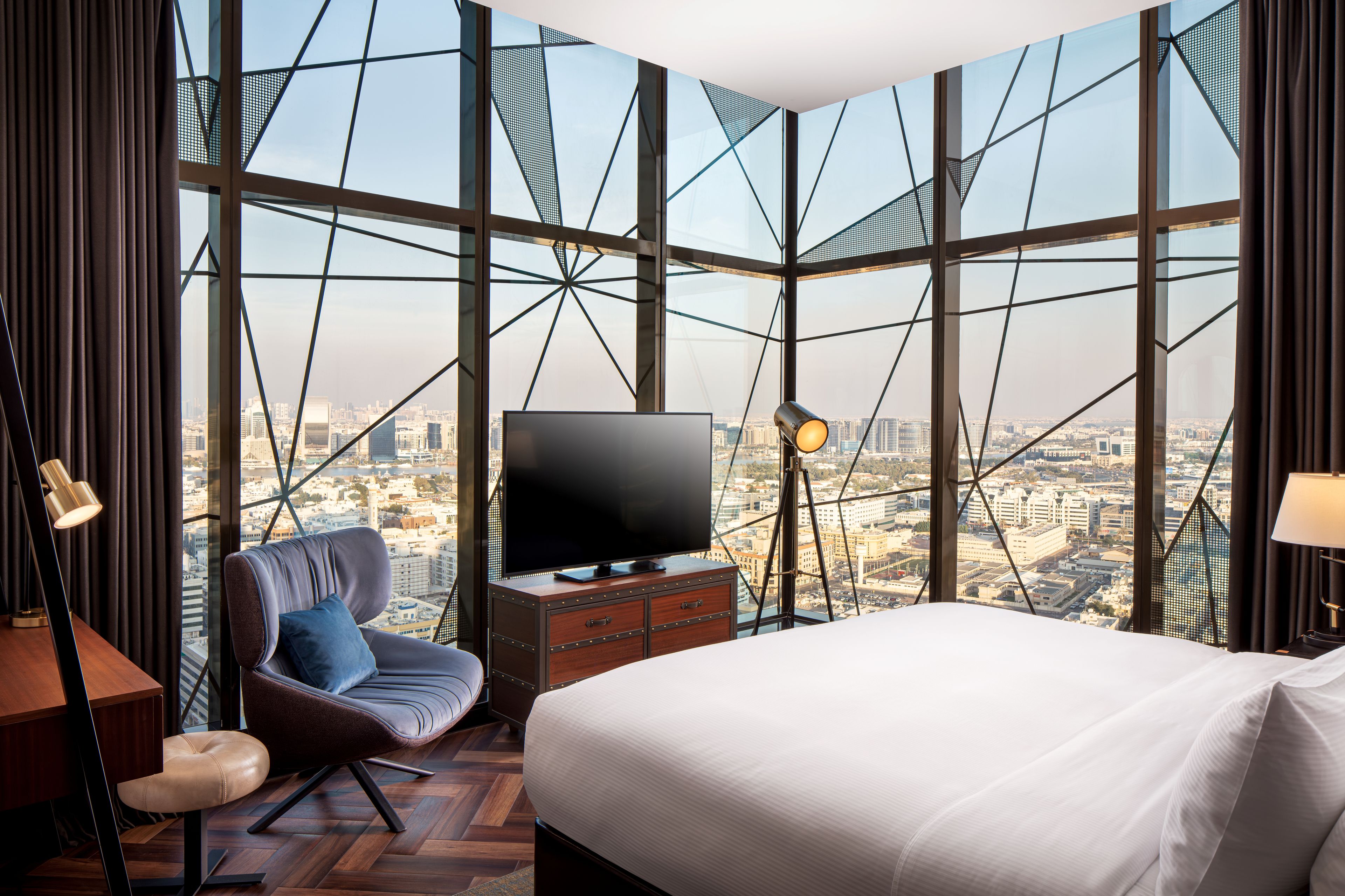 Ein modernes Hotelzimmer mit einem großen Bett, einem Fernseher, einem Sessel und Blick auf eine Skyline durch eine geometrisch gestaltete Glasfassade.
