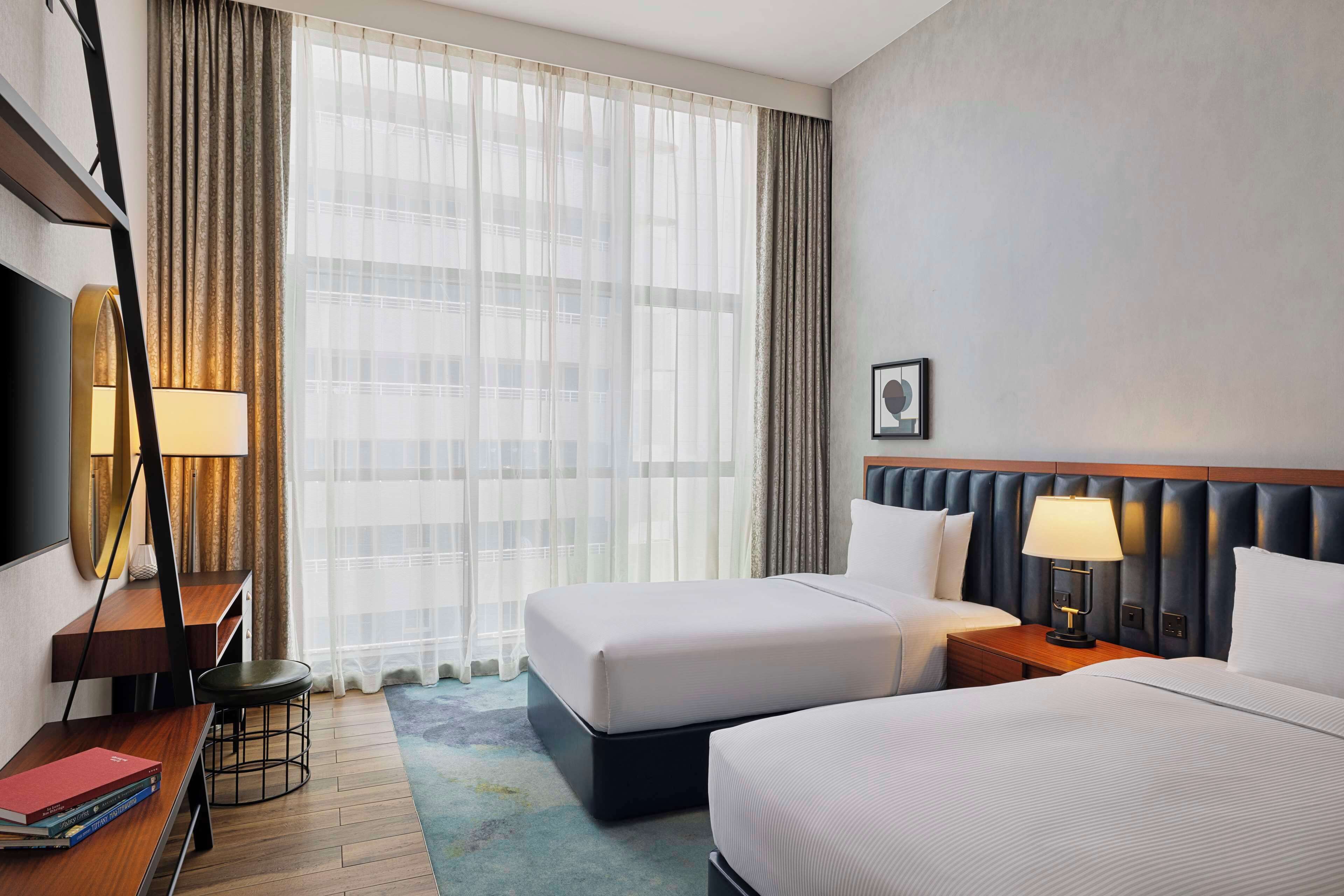 Een hotelkamer met twee bedden, een bureau met stoel, een televisie en een groot raam met gordijnen.