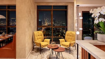 A modern hotel room with a large window offering a city view, two yellow armchairs, a small round table, and a potted orchid.