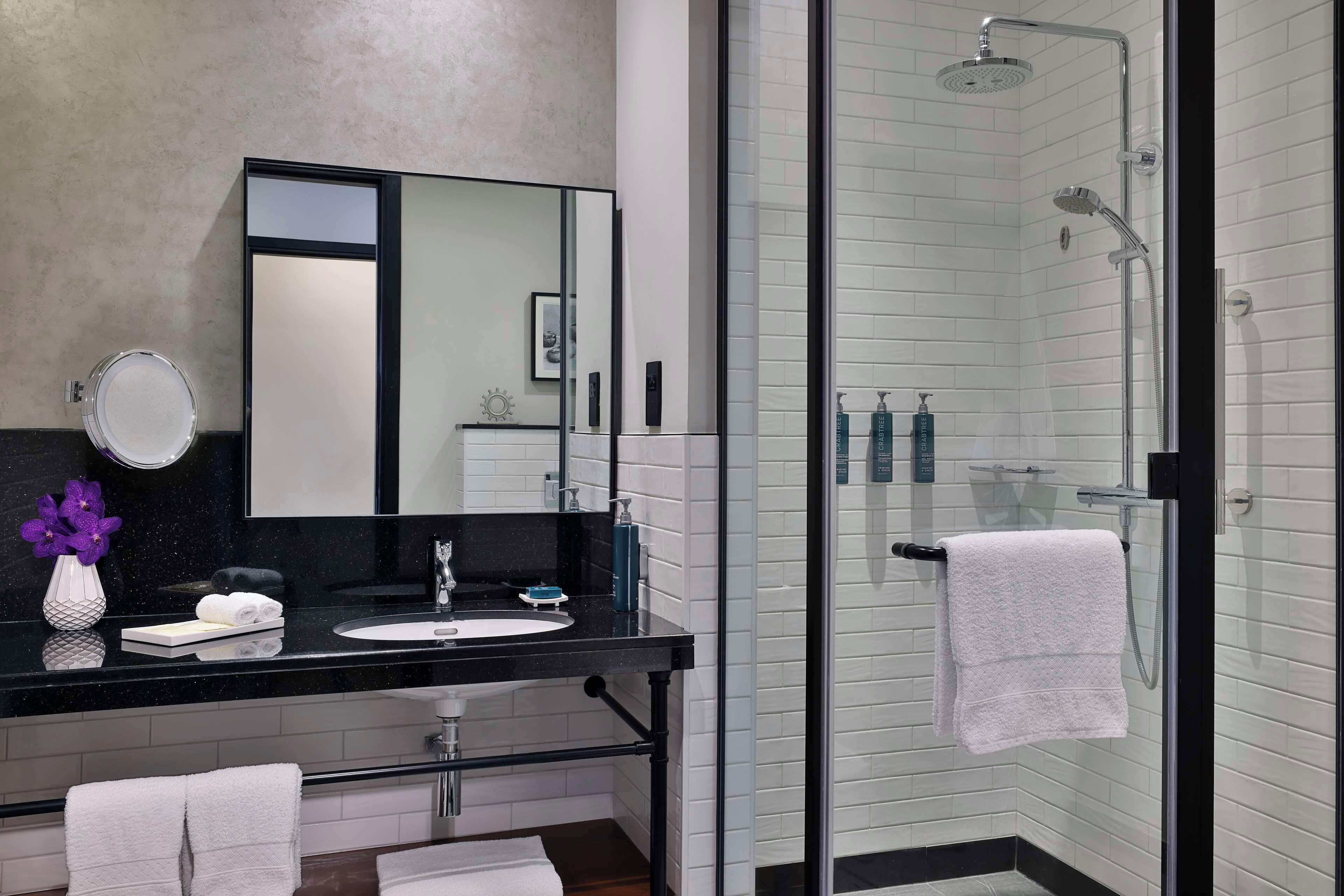 Un baño moderno con encimera negra, espejo redondo, ducha con mampara de vidrio y azulejos blancos.