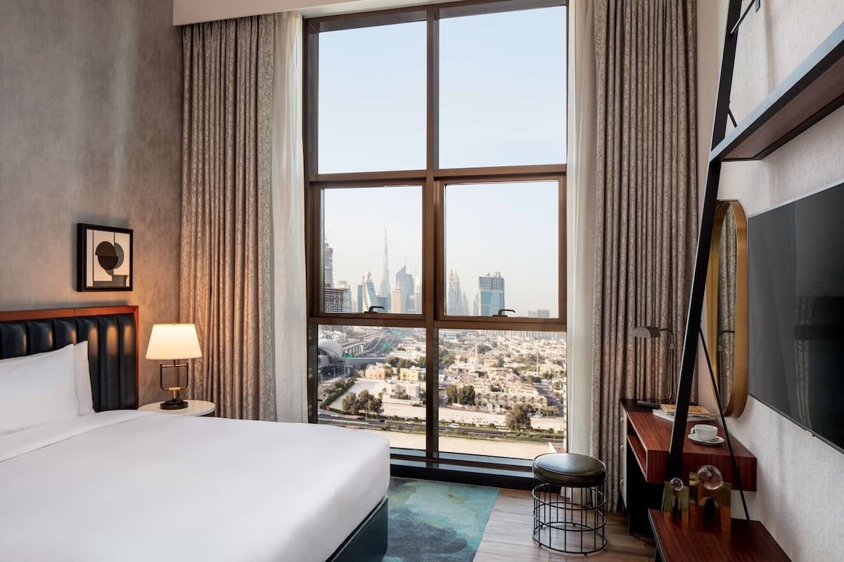 A hotel room with a large window offering a city view, a bed with white linens, a bedside table with a lamp, and a small seating area with a chair and table.