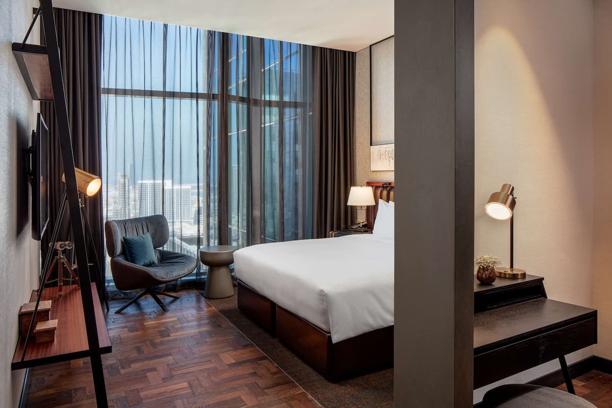 A modern hotel room with a large bed, a desk, a chair, a small table, and a large window with curtains.