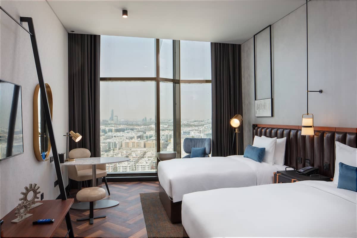 A hotel room with two beds, a desk, a chair, a small table, and a large window offering a city view.