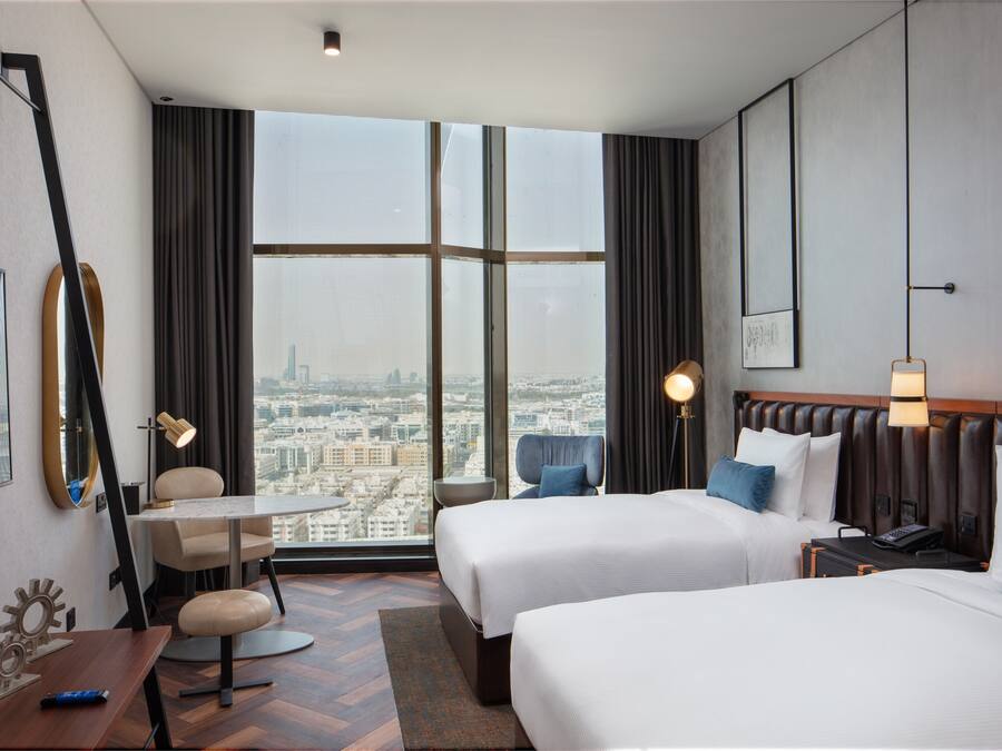 Une chambre d’hôtel avec deux lits, un bureau, une chaise, une petite table et une grande fenêtre donnant sur la ville.
