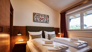 Een net opgemaakt bed met witte lakens en een opgevouwen handdoek, een houten hoofdbord, een nachtkastje met een lamp, en een raam met gordijnen.