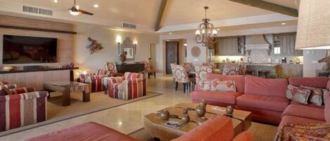 A spacious living room with a large sofa set, a coffee table, and a television. There is a kitchen area in the background with cabinets and a countertop.