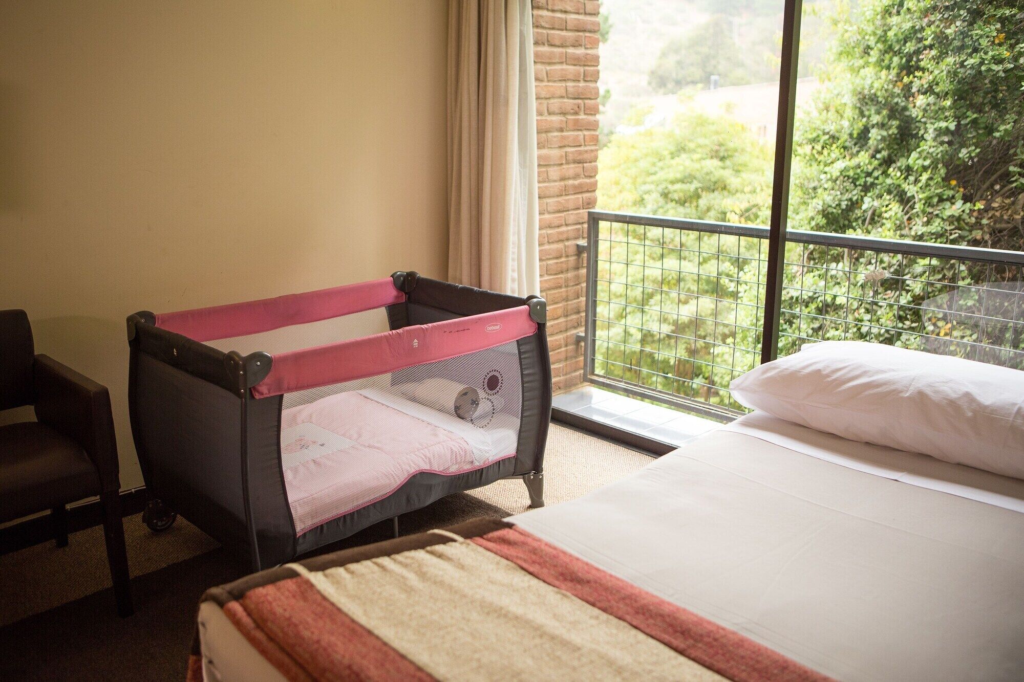 A room with a bed, a crib, and a view of greenery through a window.