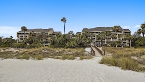 A beachfront hotel with multiple floors, palm trees, and a sandy beach in the foreground.