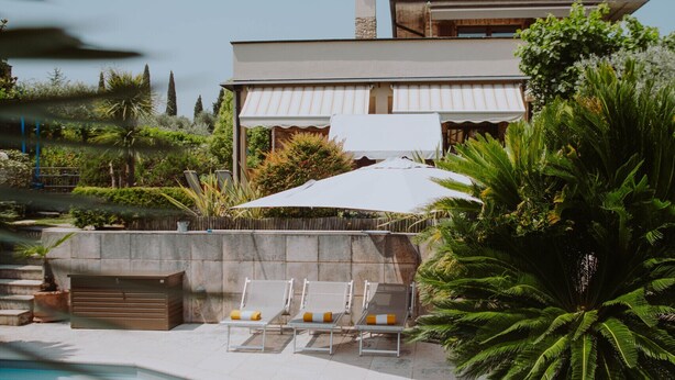 A pool area with lounge chairs, a large umbrella, and a building with chimneys. - Villa Riai (Moniga del Garda)