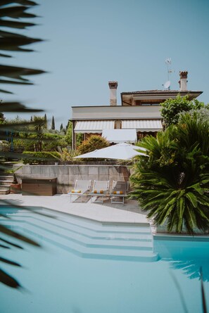Zona de piscina con sillas de descanso, una sombrilla grande y un edificio con chimeneas.
