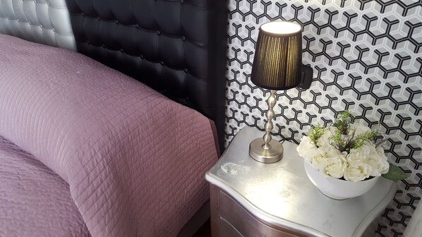 A neatly made bed with a pink quilt, a black tufted headboard, a modern lamp, and a white vase with flowers on a bedside table. - Chez Alice Vatican (Rome)