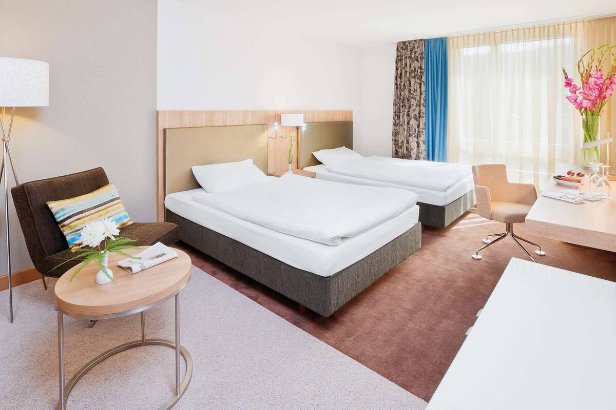 Een hotelkamer met twee bedden, een bureau, een stoel, een lamp en een vaas met bloemen.