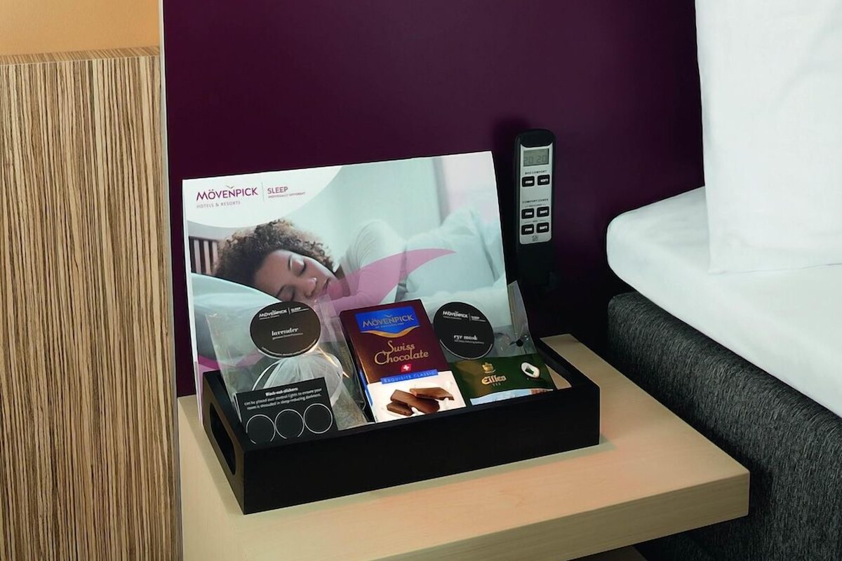 Habitació d'hotel amb una taula de nit on es veu una revista sobre el son, un comandament a distància i una caixa de xocolata suïssa.