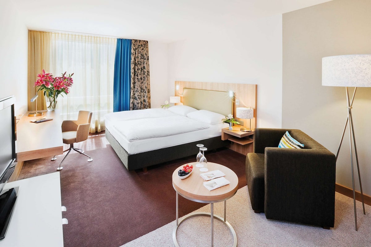 Een hotelkamer met een bed, nachtkastje, lamp, bureau, stoel en een klein rond tafeltje met een schaal fruit.