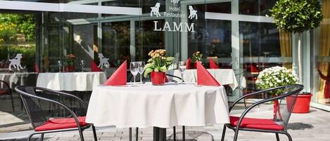 Un área de comedor al aire libre con mesas dispuestas para dos, con sillas rojas y manteles blancos.