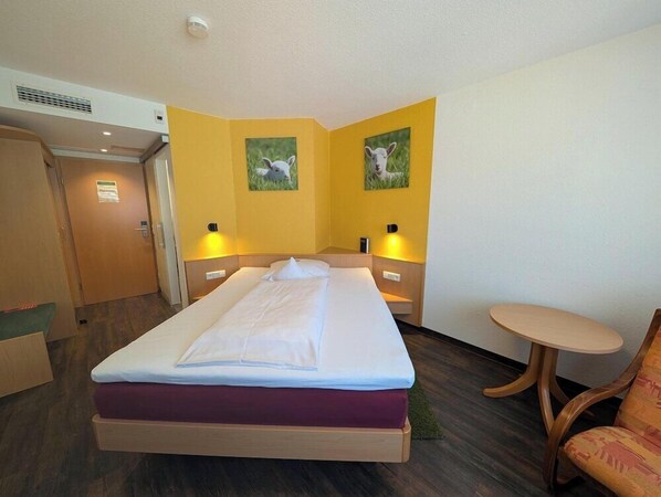 Una habitación de hotel con cama, mesitas de noche, una silla y dos lámparas fijadas a la pared.