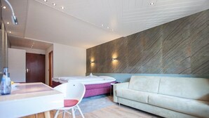 A modern hotel room with a wooden floor, a white sofa, a pink chair, a small table, and a large wall-mounted TV.