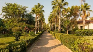 A paved pathway lined with palm trees and well-maintained hedges, leading towards a building with a red-tiled roof. - Perre Delta Hotel (Alanya)