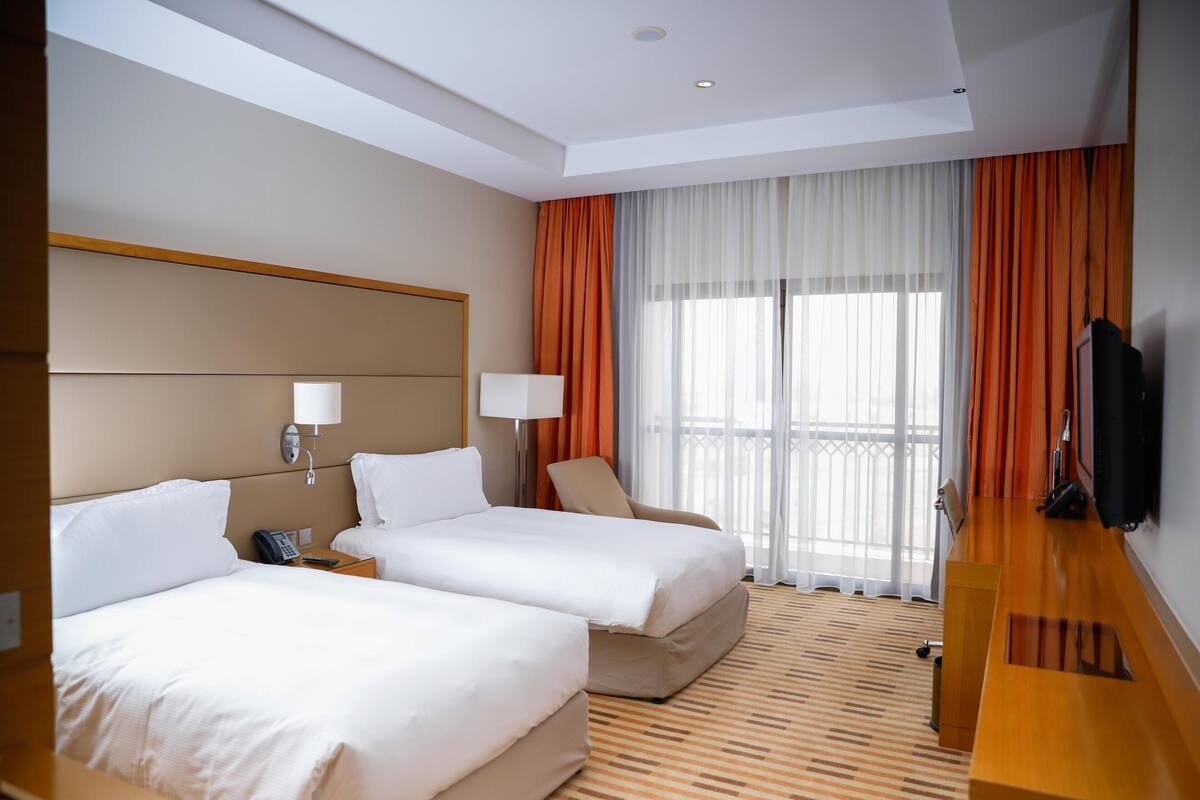 A hotel room with two beds, a TV, a desk, and a balcony with curtains.