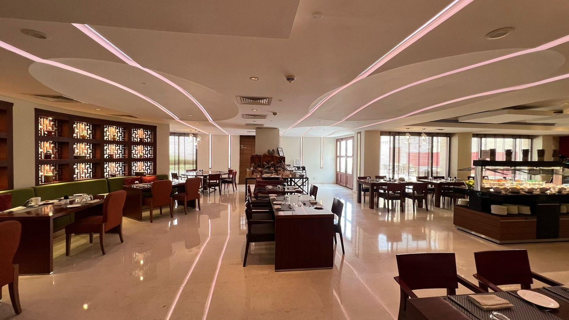 A spacious hotel dining area with multiple tables and chairs, a central seating area, and a buffet counter.