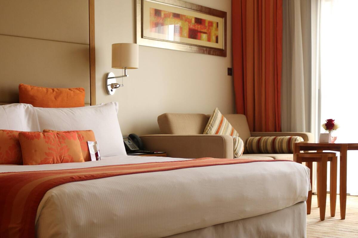 A hotel room with a large bed, a chair, a small table, a lamp, and a painting on the wall.