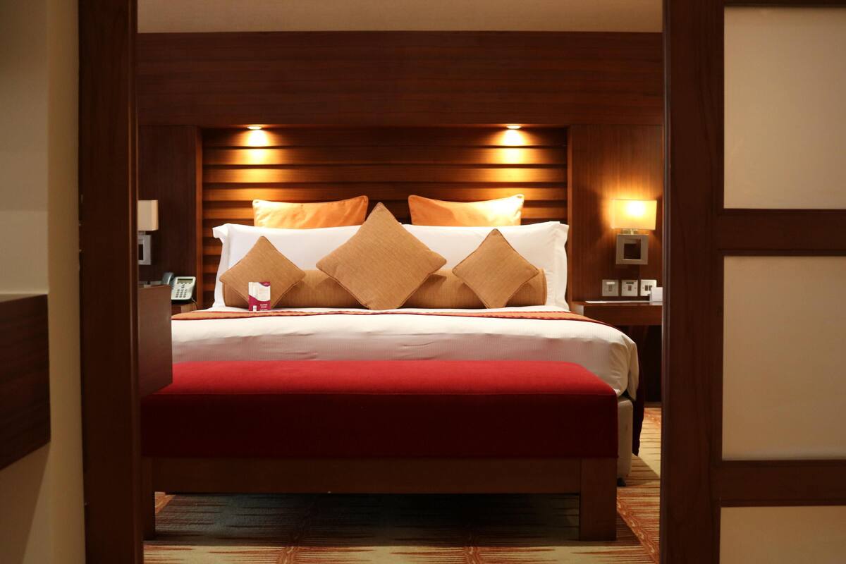 A hotel room with a large bed, wooden headboard, bedside lamps, and a red accent pillow.