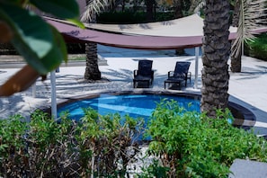 Piscina com duas espreguiçadeiras, rodeada por palmeiras e uma área com sombra.