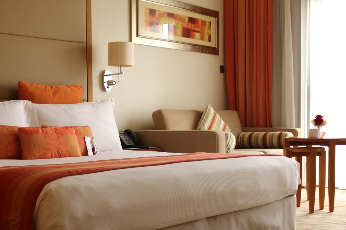 Habitación de hotel con una cama grande, una silla, una mesita, una lámpara y un cuadro en la pared.