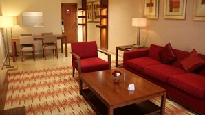 Habitación de hotel con sofá rojo, una mesita de centro de madera, un área de comedor con mesa y sillas, y un televisor.