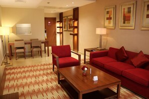Habitación de hotel con sofá rojo, una mesita de centro de madera, un área de comedor con mesa y sillas, y un televisor.